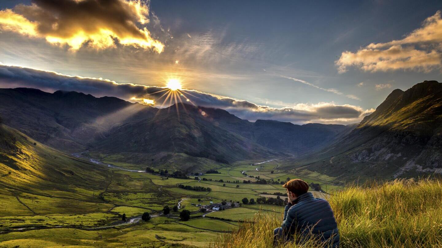 Man sitting in long grass on the side of a fell watching the sunset