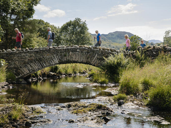 Un grupo de personas camina sobre un puente de piedra sobre un río, rodeado de paisaje verde y colinas bajo un cielo soleado.