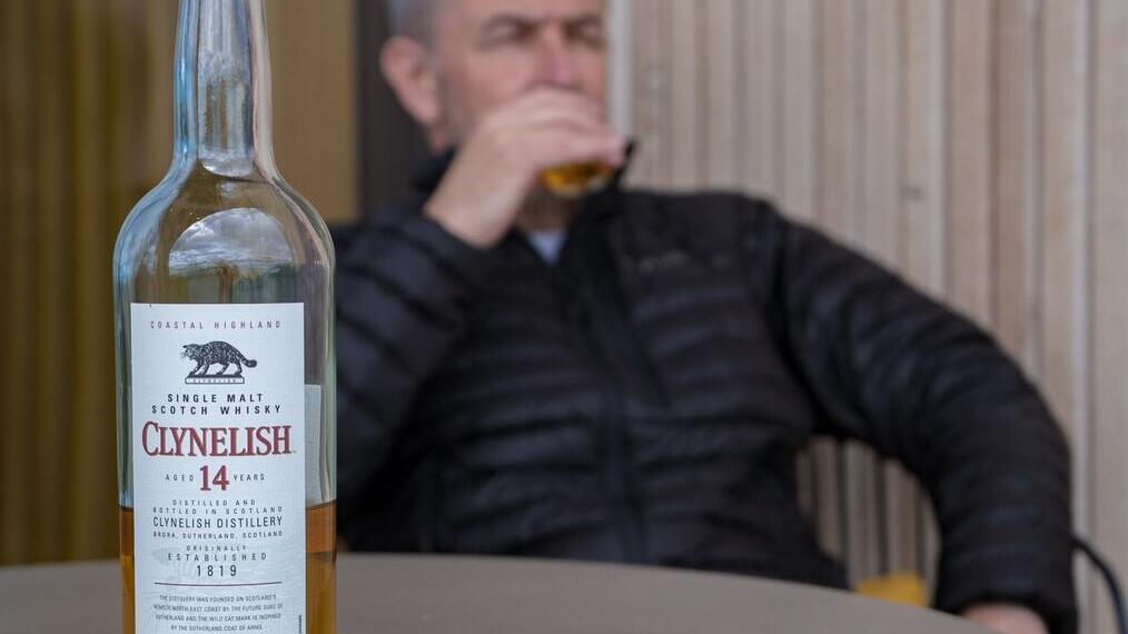 Man sitting at table drinking a glass of whisky with a bottle of whisky in the foreground