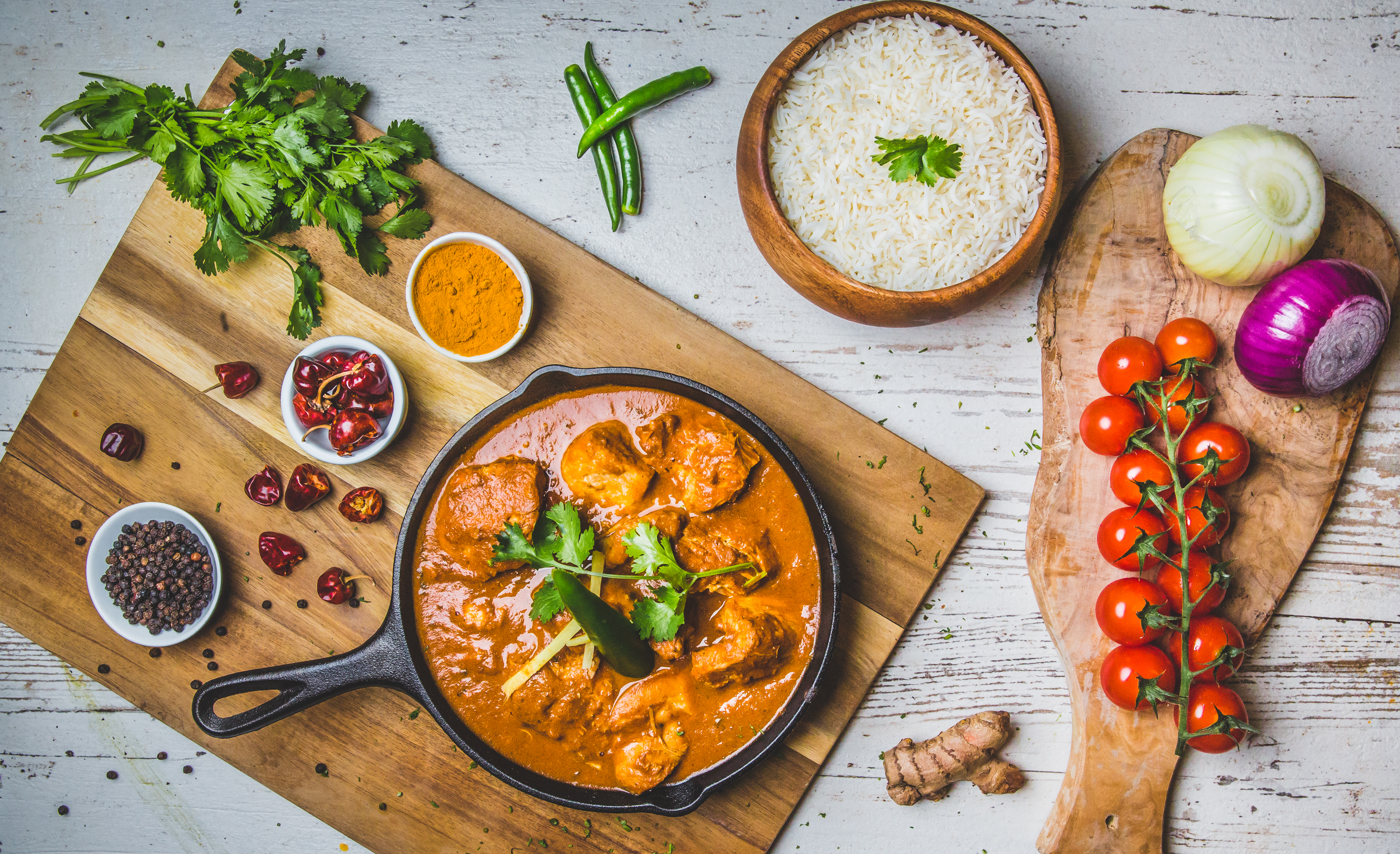 A skillet of chicken curry with rice, surrounded by fresh vegetables and spices on a rustic table.