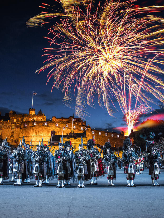 The arena at the Edinburgh Military Tattoo during a performance of the military event, parade ground and packed spectator seating. A light show projecting onto the castle walls. Marching band with a leading conductor, and massed pipers playing the bagpipes. Fireworks exploding in the night sky.
