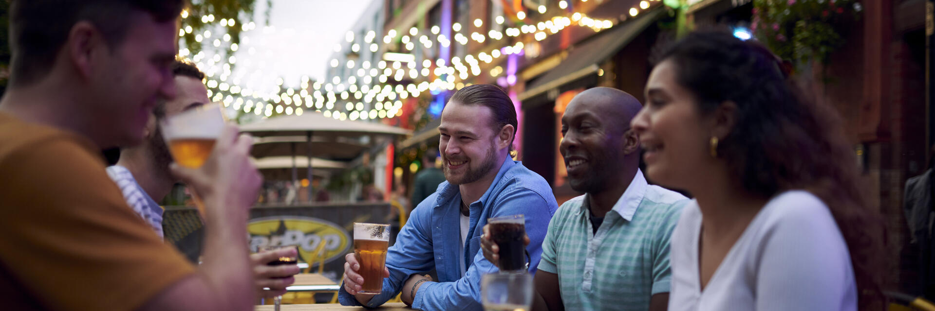 Friends having a drink sat at an outside table of a bar with fairy lights hanging behind