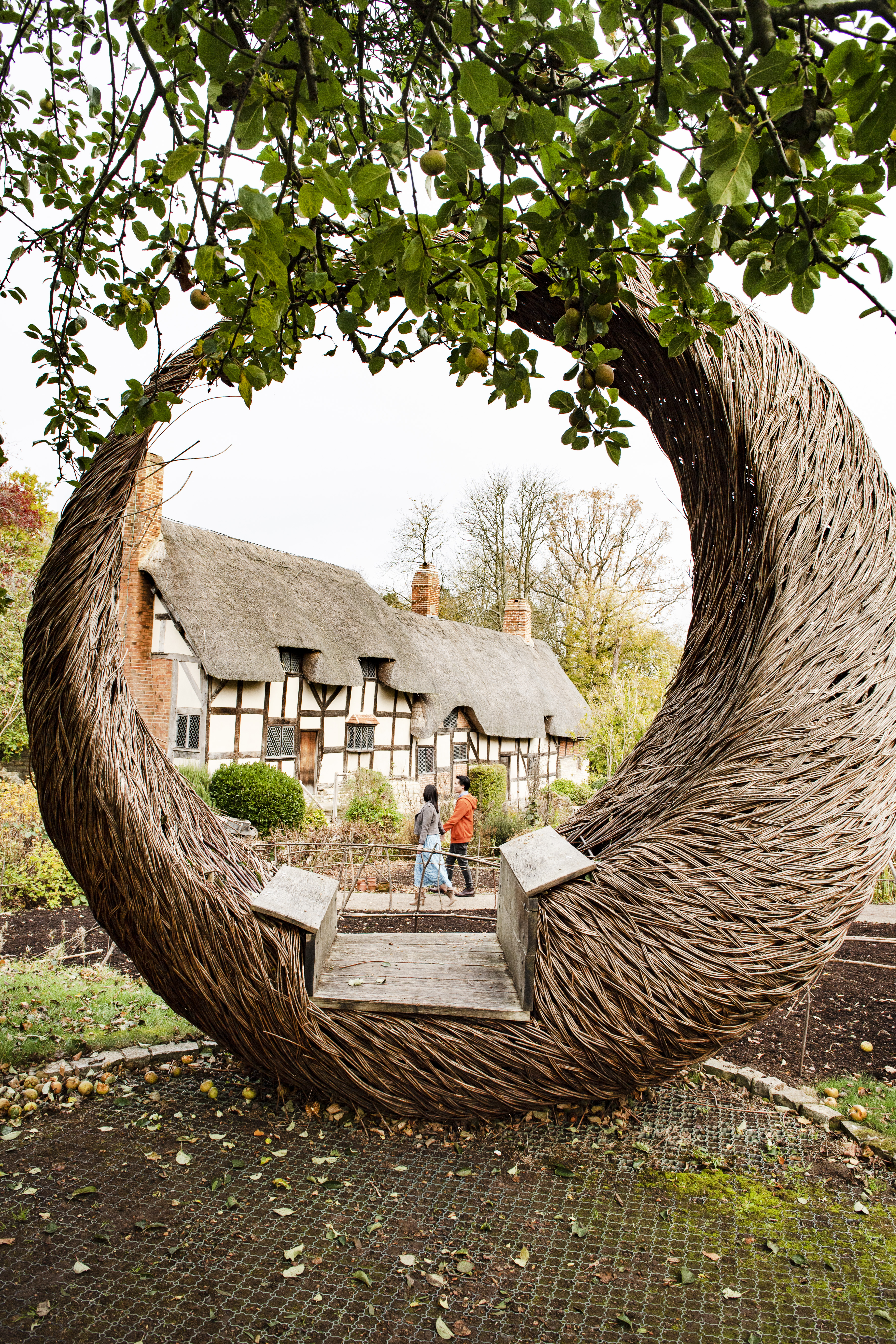 Couple walking near a cottage seen through a large willow crescent shaped sculpture in a garden
