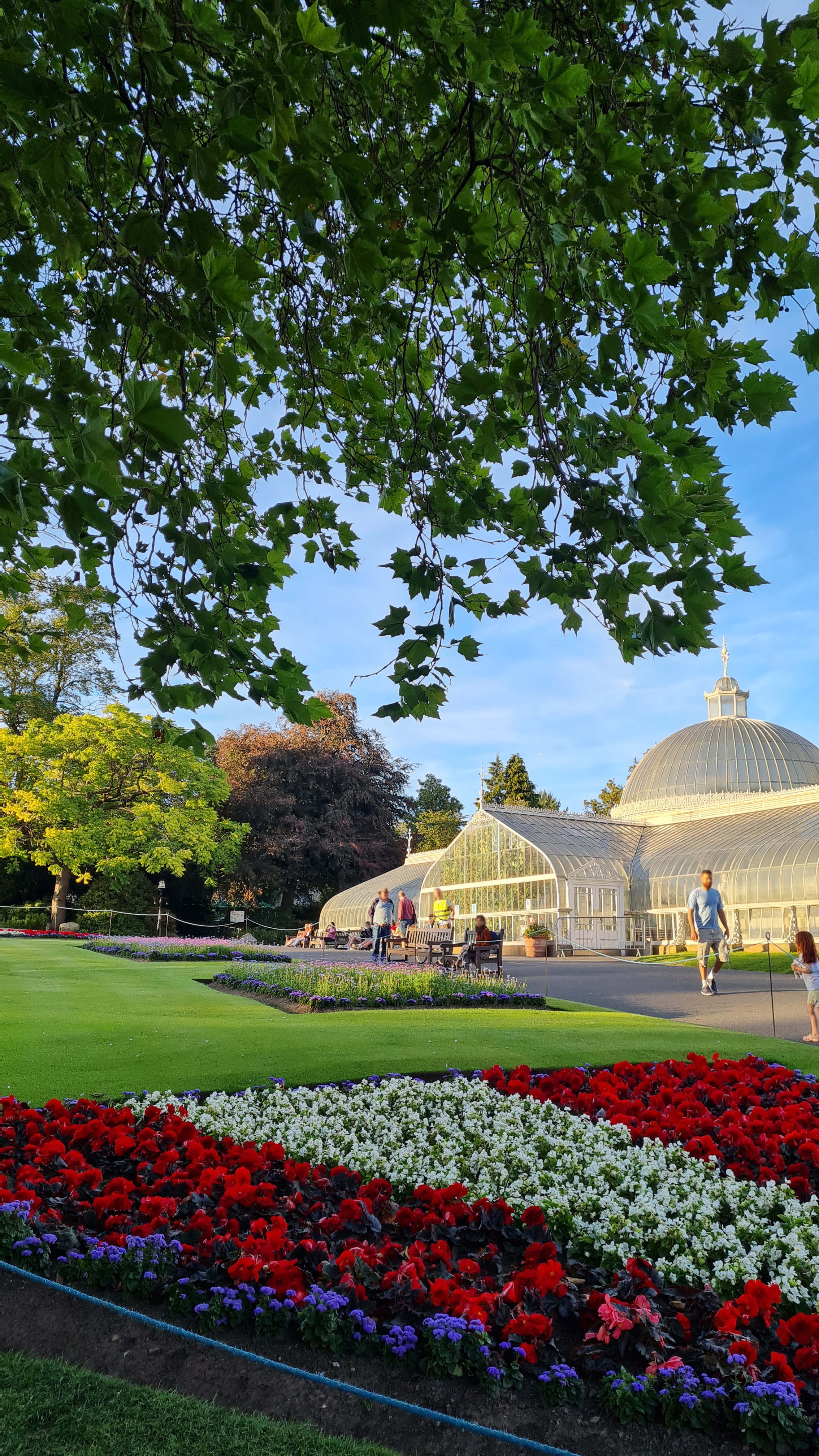 Large domed glass house building in the background in the sunshine, with people outside walking though the park, flower beds in foreground.