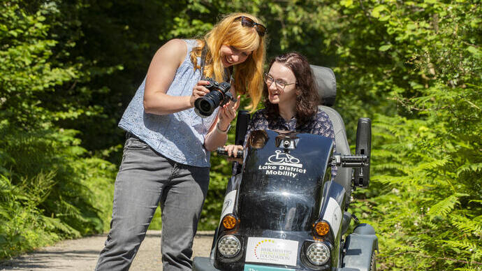 A woman stands holding a camera showing the pictures to a woman who sits in a tramper