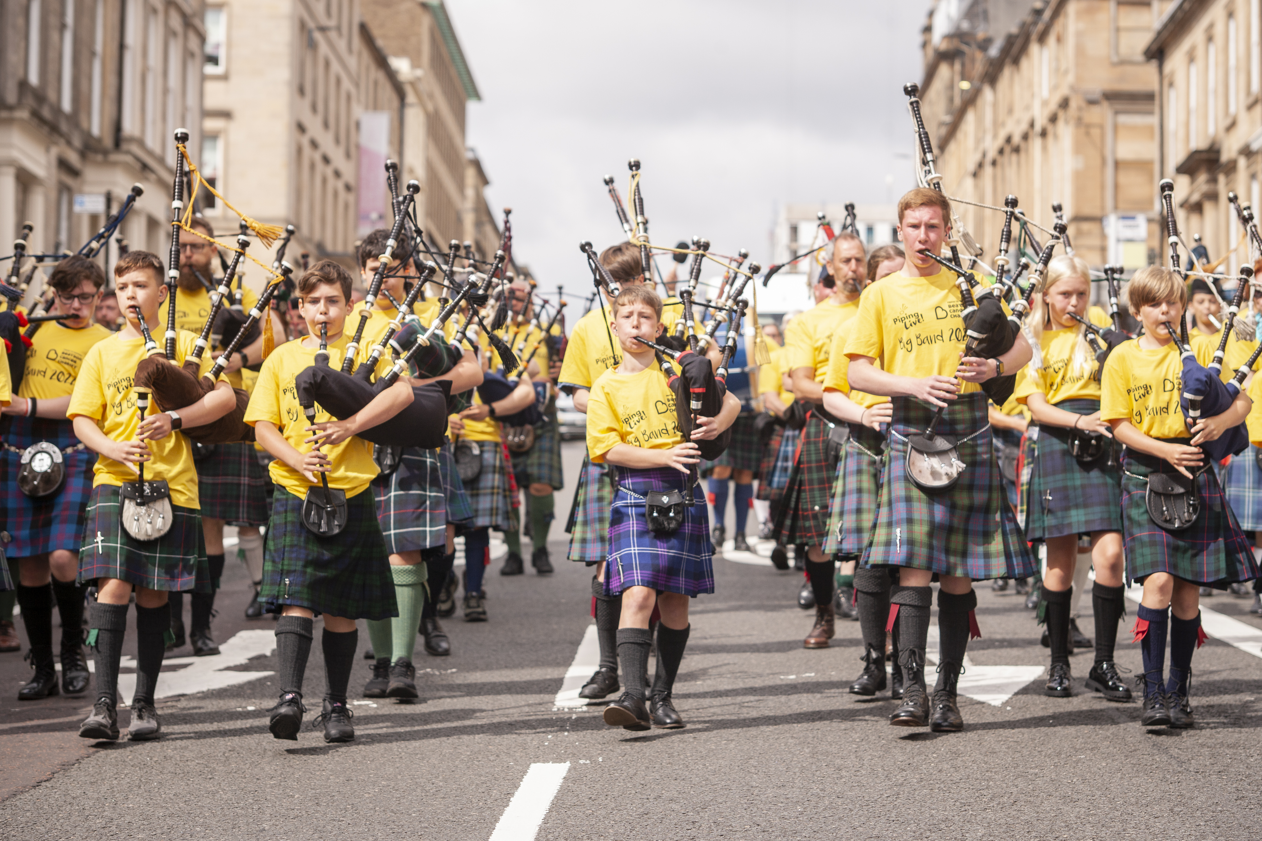 Children's pipping band wearing kilts and yellow T-shirts play the bagpipes while marching down the street.