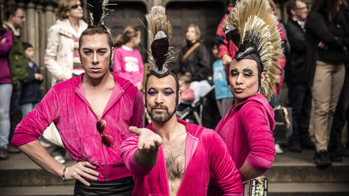 Tre persone con trucco drammatico e creste mohawk in abiti rosa posano con sicurezza a un evento pubblico con una folla sullo sfondo.