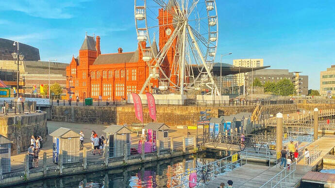 Ferris wheel and historic red-brick building by waterfront, with people walking on docks and clear blue sky above.