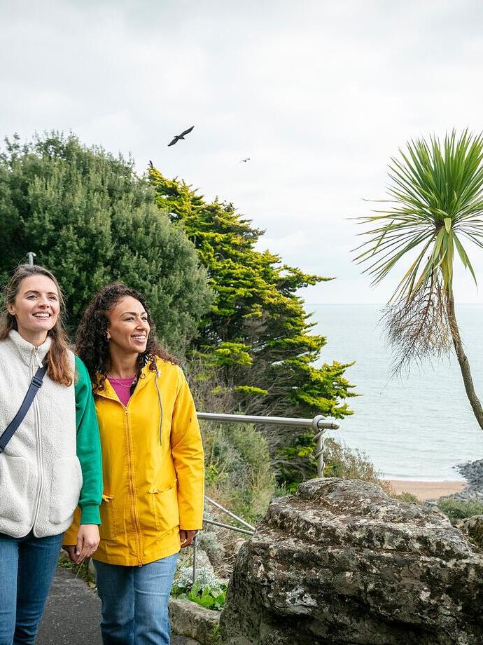 Two people walk on a scenic coastal path lined with greenery and palm trees, overlooking the sea on a cloudy day.