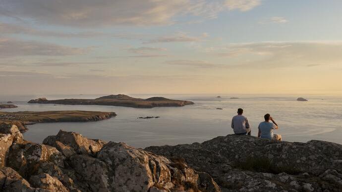 Two men sat on a rock cliff looking out to sea at dusk
