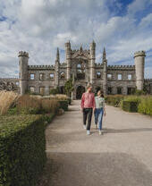 A couple walks down a path toward a large castle ruin with towers and arched windows, surrounded by gardens and dramatic clouds.