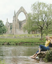 A man and a woman sit by a river near a heritage Abbey