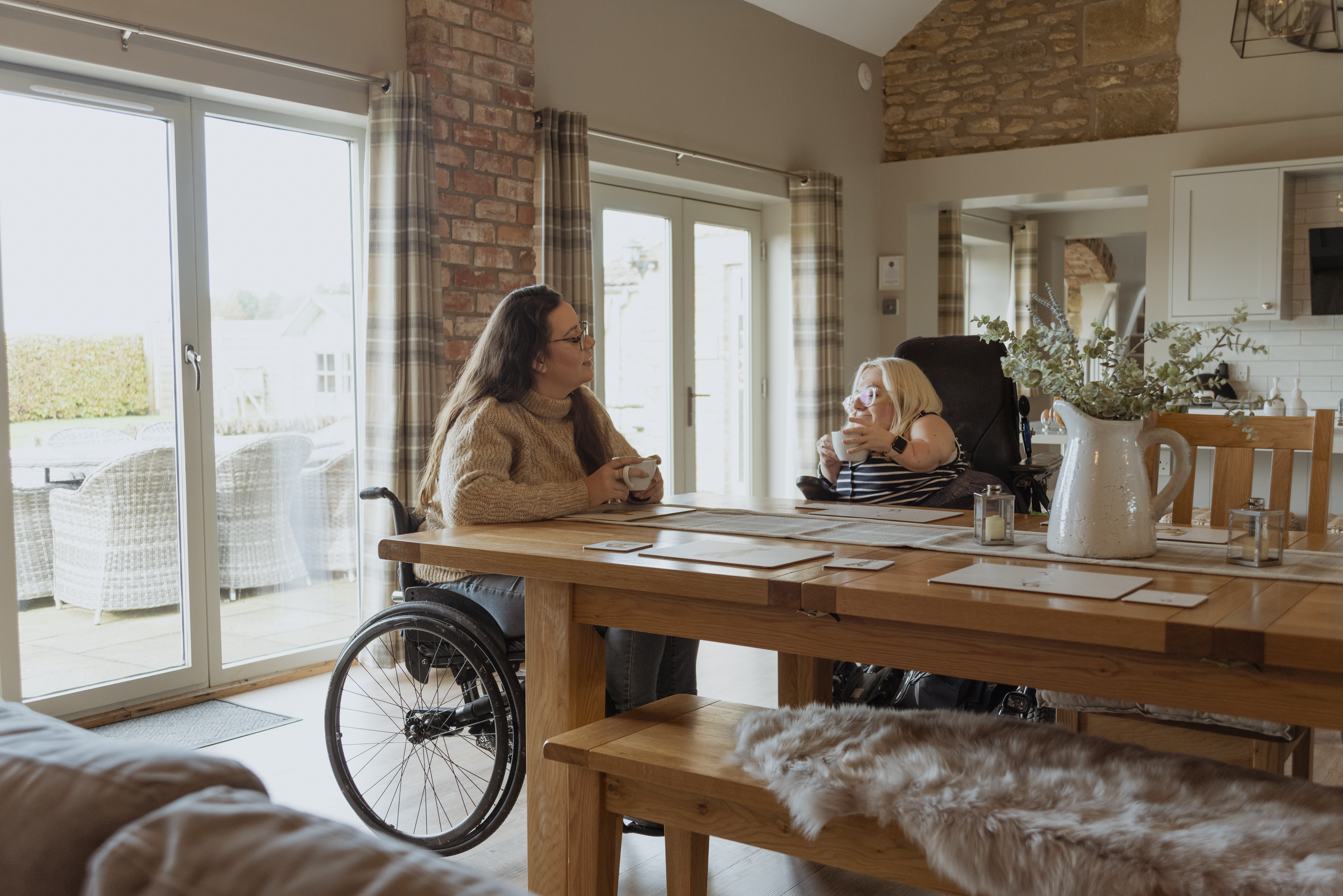 Two friends having a cup of tea at a holiday cottage. One is in a mobility scooter, the other in a wheelchair.