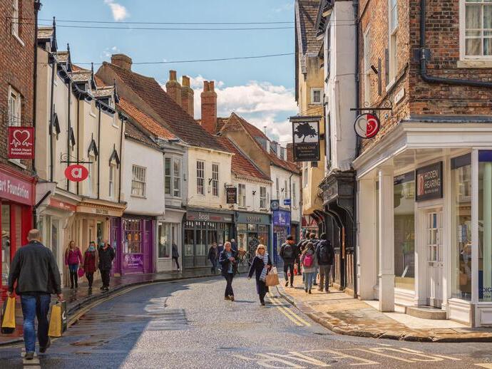Shopping street just after a rain shower. Historic buildings line the street and shoppers walk between them. A sky with cloud is overhead.