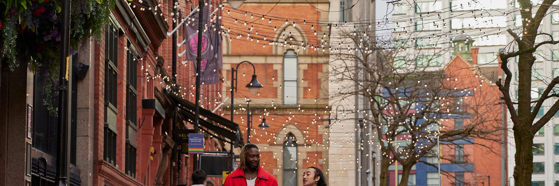 Rue pavée avec terrasses de cafés, guirlandes lumineuses suspendues et passants, bâtiments historiques et modernes en arrière-plan.