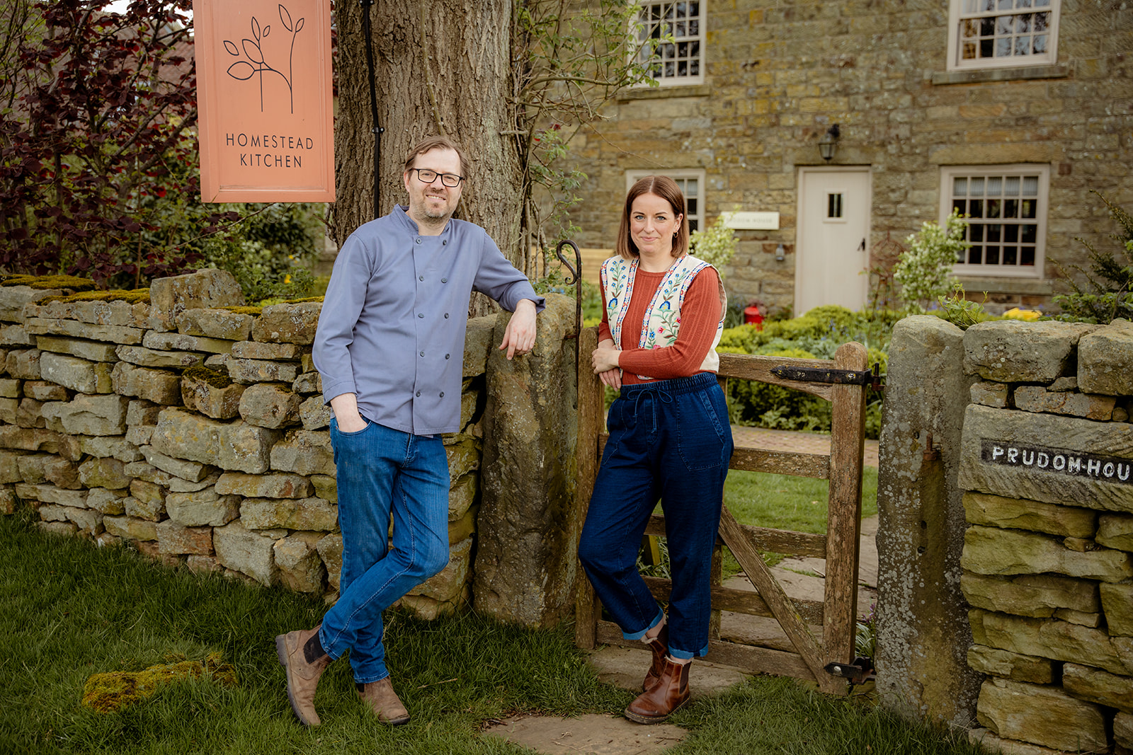 Deux personnes debout devant un cottage à la campagne. Une pancarte indique : « Homestead Kitchen »