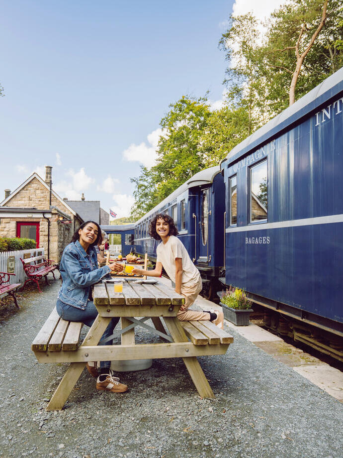 Two people dining at a picnic table on a historic train station platform with a blue train and Bassenthwaite Lake station sign visible.