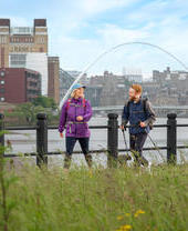 A woman and a man walk along a riverside with iconic buildings and a bridge beyond