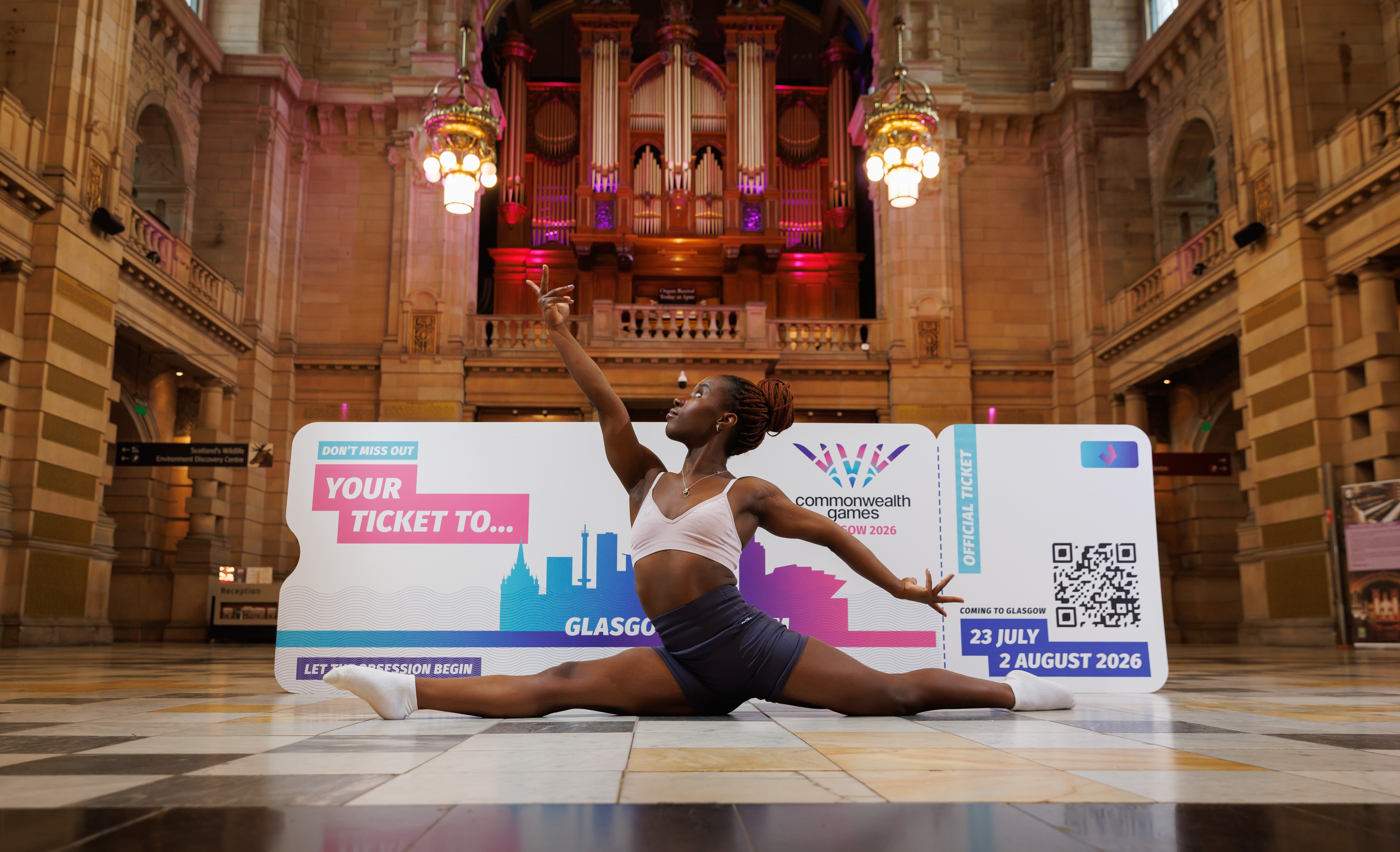 A dancer in athletic wear performs a split pose in the foyer of an ornate building, with a sign promoting the Commonwealth Games Glasgow 2026 in the background.