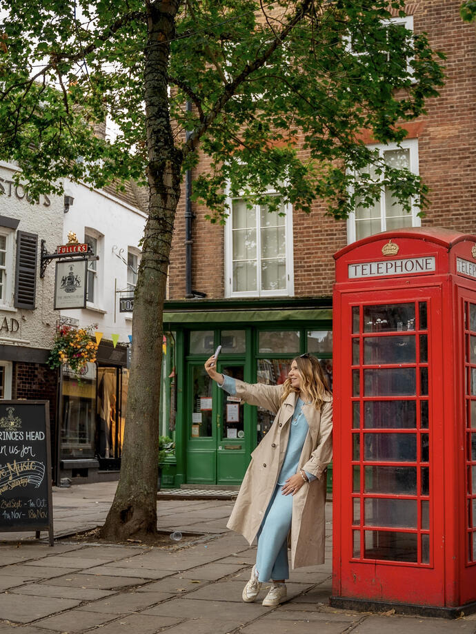 A woman leaning against a red telephone box and takes a selfie
