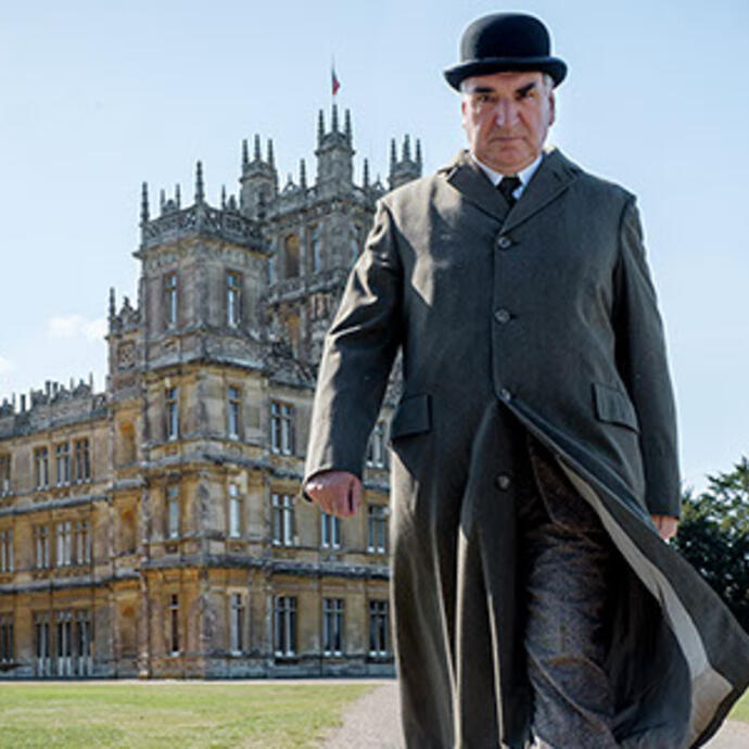 A person in period clothing walks in front of a grand historic mansion with ornate architecture and landscaped grounds.