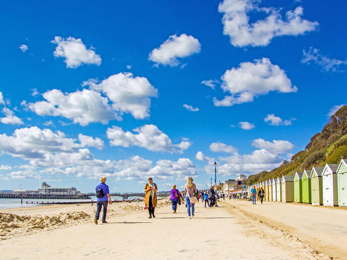 People walking along the coast next to colourful beach huts on a sunny day.