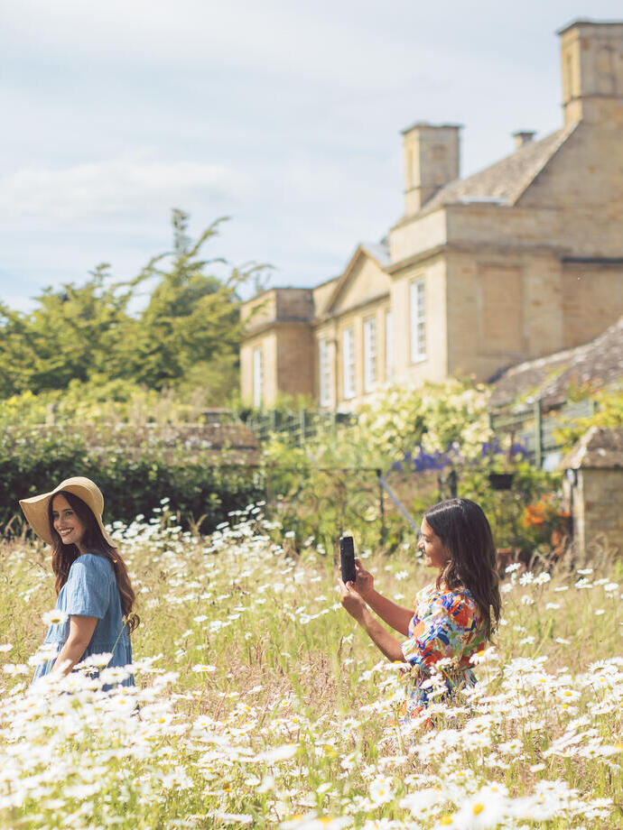 Two women enjoy gardens full of daisies in front of a stone house