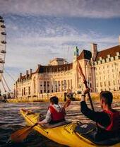 Dos personas en kayak en el río Támesis con el London Eye y un edificio histórico al fondo.