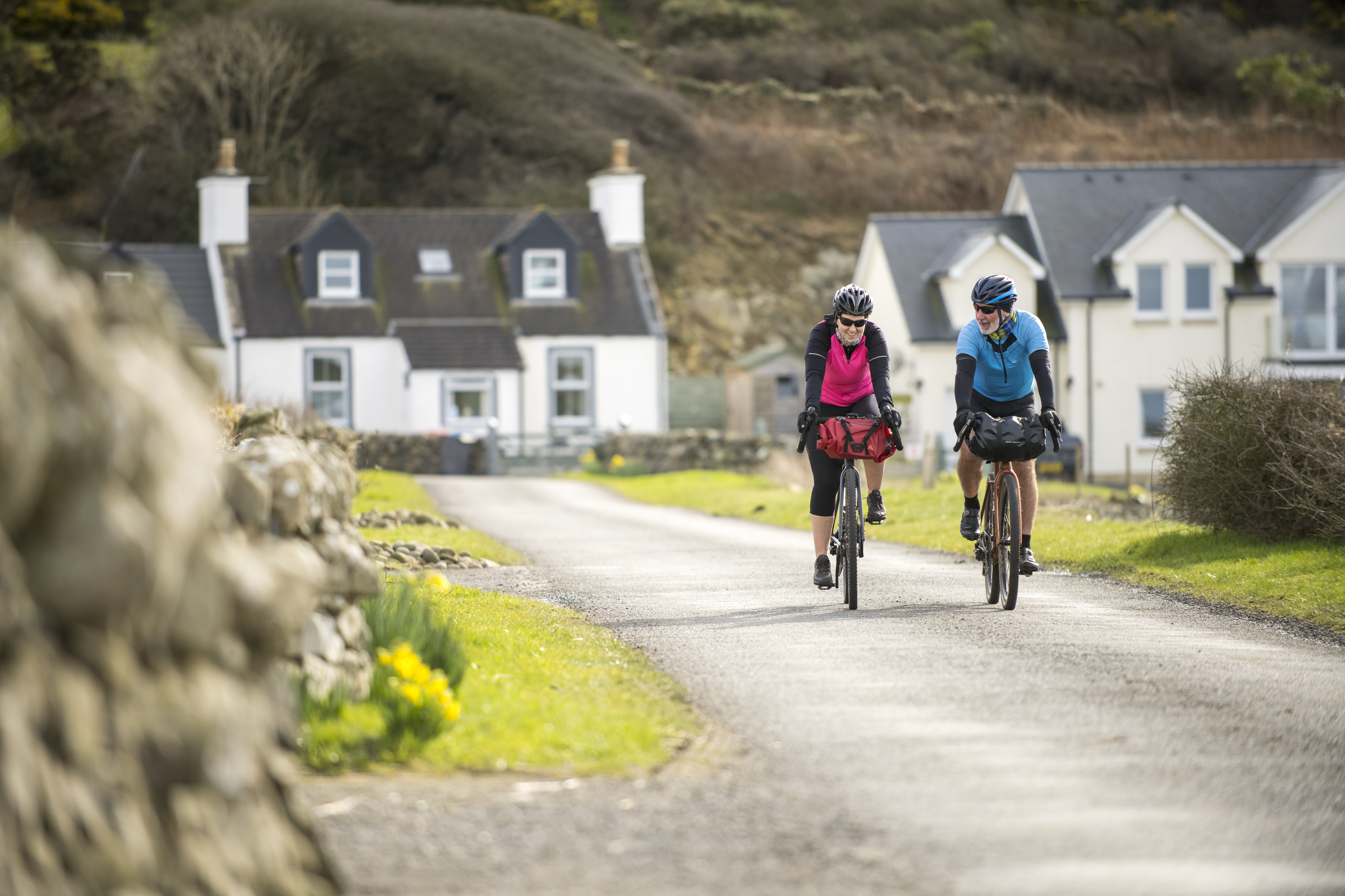 Two cyclists ride on a rural lane past stone walls and houses, with green grass and hills in the background.