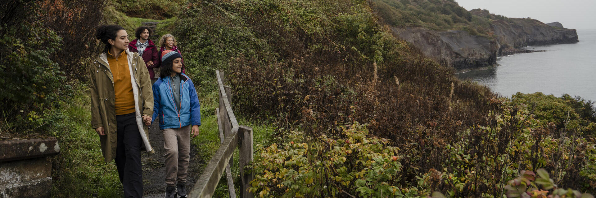 Group walking along a coastal path with lush greenery, cliffs, and sea in the background on a cloudy day.