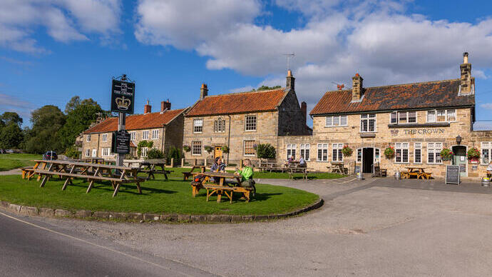 Stone buildings with red tile roofs, outdoor pub tables, and people dining on a village green under a blue sky.