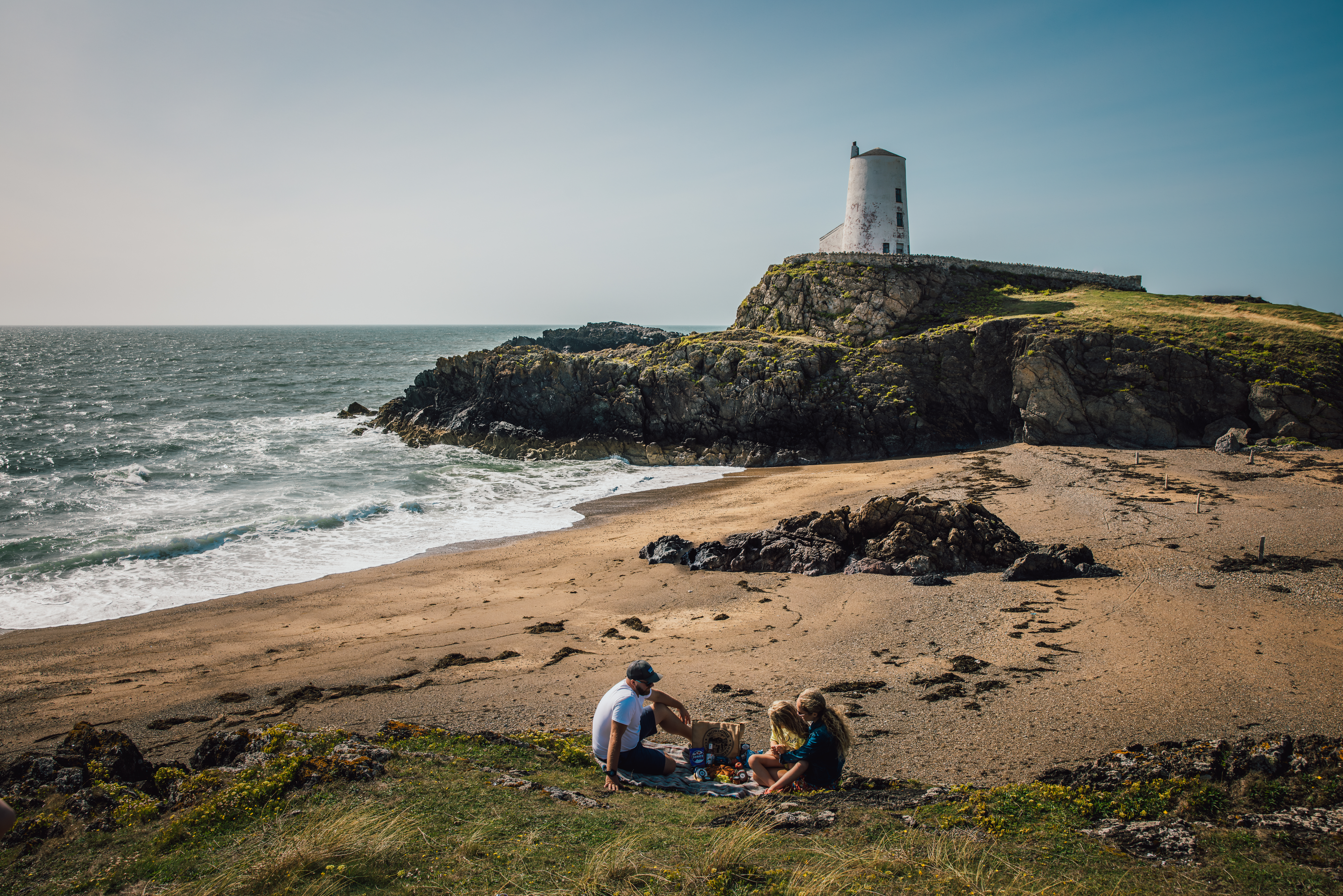 Une jeune famille pique-niquant sur une plage avec un phare en arrière-plan.