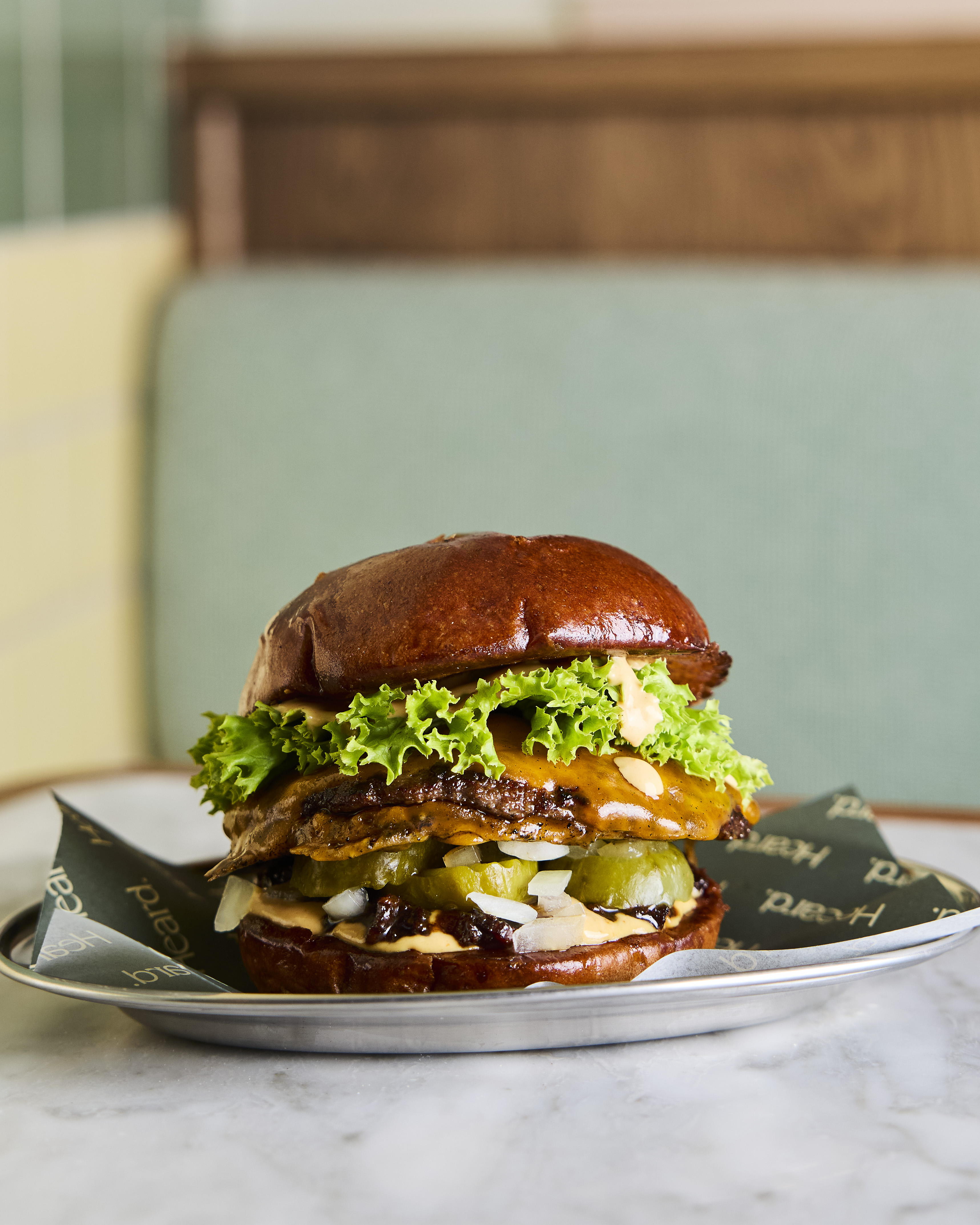 Close-up of a gourmet burger with lettuce, cheese, pickles, onions, and sauce in a glossy brioche bun on a plate in a restaurant setting.