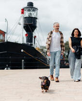 Two people walking a small dog along a paved waterfront with a historic ship and residential buildings in the background.