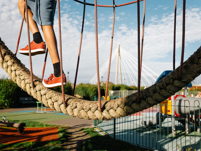 A child wearing red shoes and shorts uses a rope play structure at a playground, with a cable-stayed bridge in the background.