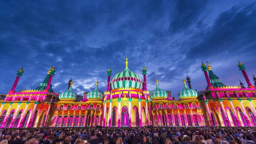 Crowd sitting on the ground in front of a large ornate building lit up with bright colours