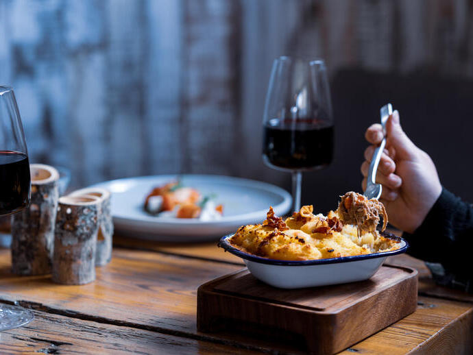 View of arm eating a mashed potatoes topped dish with two glasses of red wine and another dish of food in soft focus in the background 