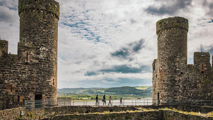 Castillo histórico con dos grandes torres y vista rural al fondo. Personas caminan por un puente entre las torres.