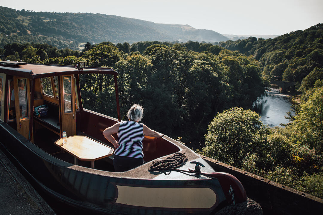 A person on a canal boat looking out over a lush green valley with a river and hills in the background.