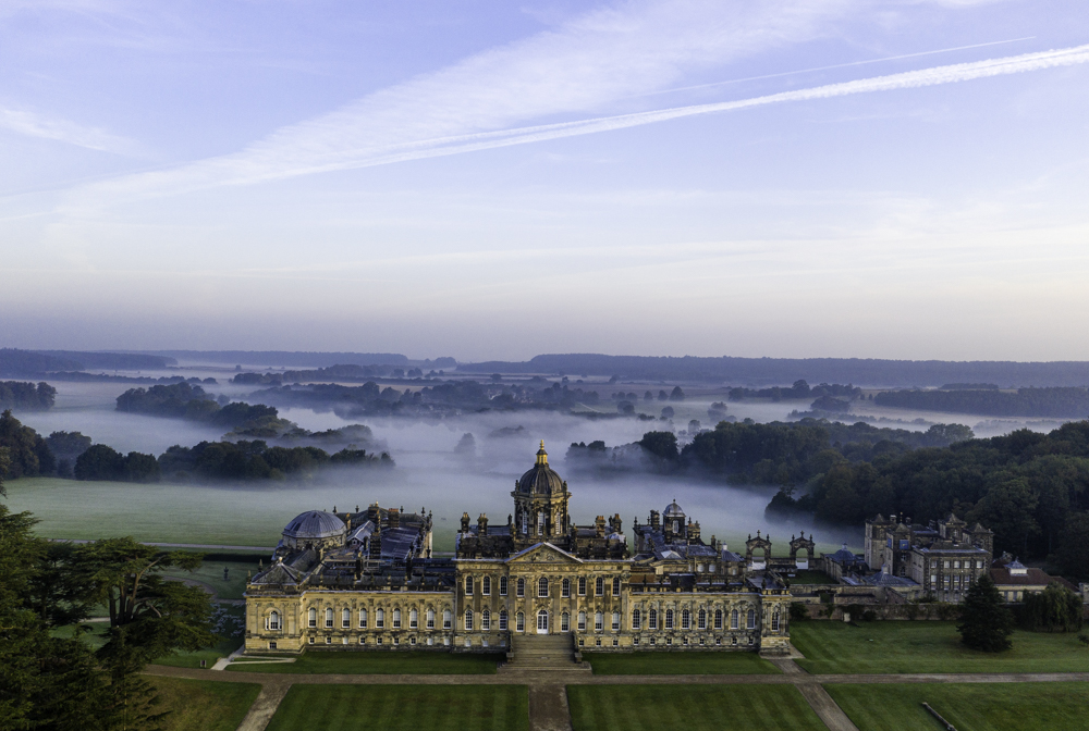Grande maison de campagne vue d'en haut avec des jardins à la française et une campagne boisée derrière, recouverte de nuages bas