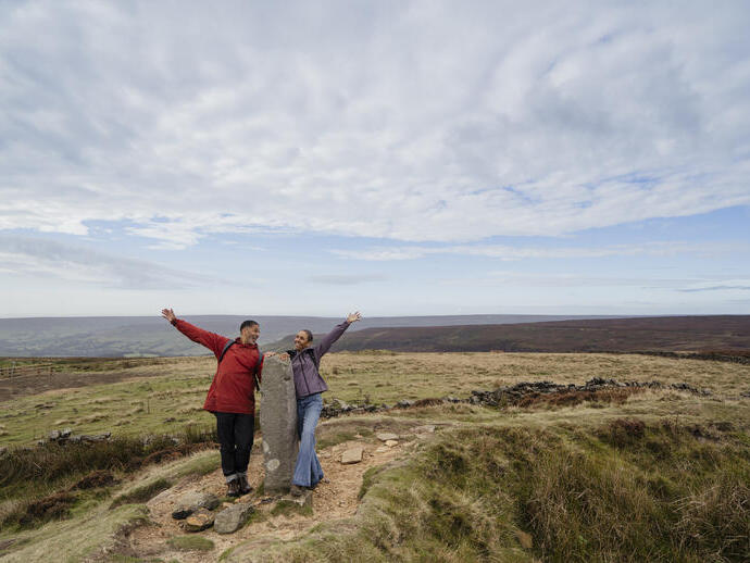 Dos personas de excursión en una zona rural abierta, junto a una piedra alta, con los brazos levantados. Al fondo, colinas y un paisaje extenso.