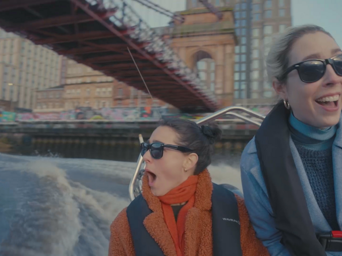 Twee vrouwen op een motorboot op een rivier, met een brug en gebouwen op de achtergrond