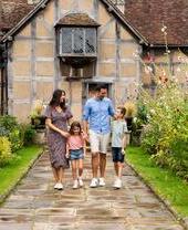 Eine Familie geht durch einen Garten vor einem historischen Haus mit schrägem Dach und blühendem Garten.