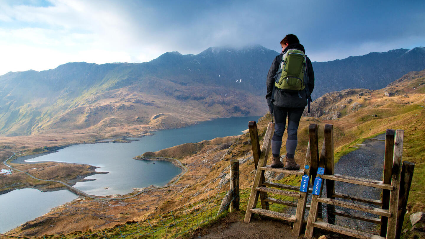Wanderer auf einem hölzernen Aussichtspunkt mit Blick auf eine weitläufige Bergkette und ein See-Panorama
