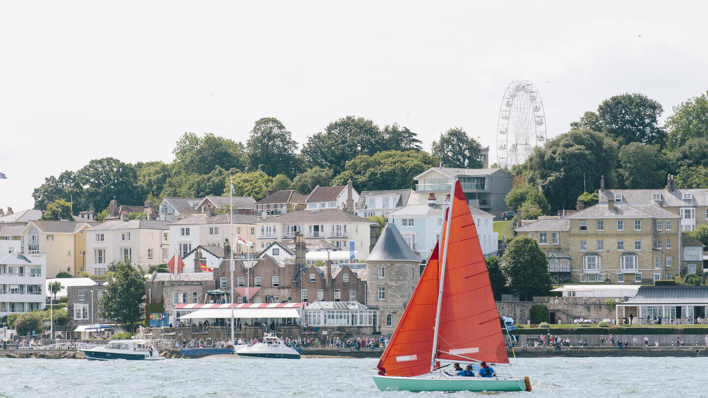Red sailing boat sailing in front of a coastal town