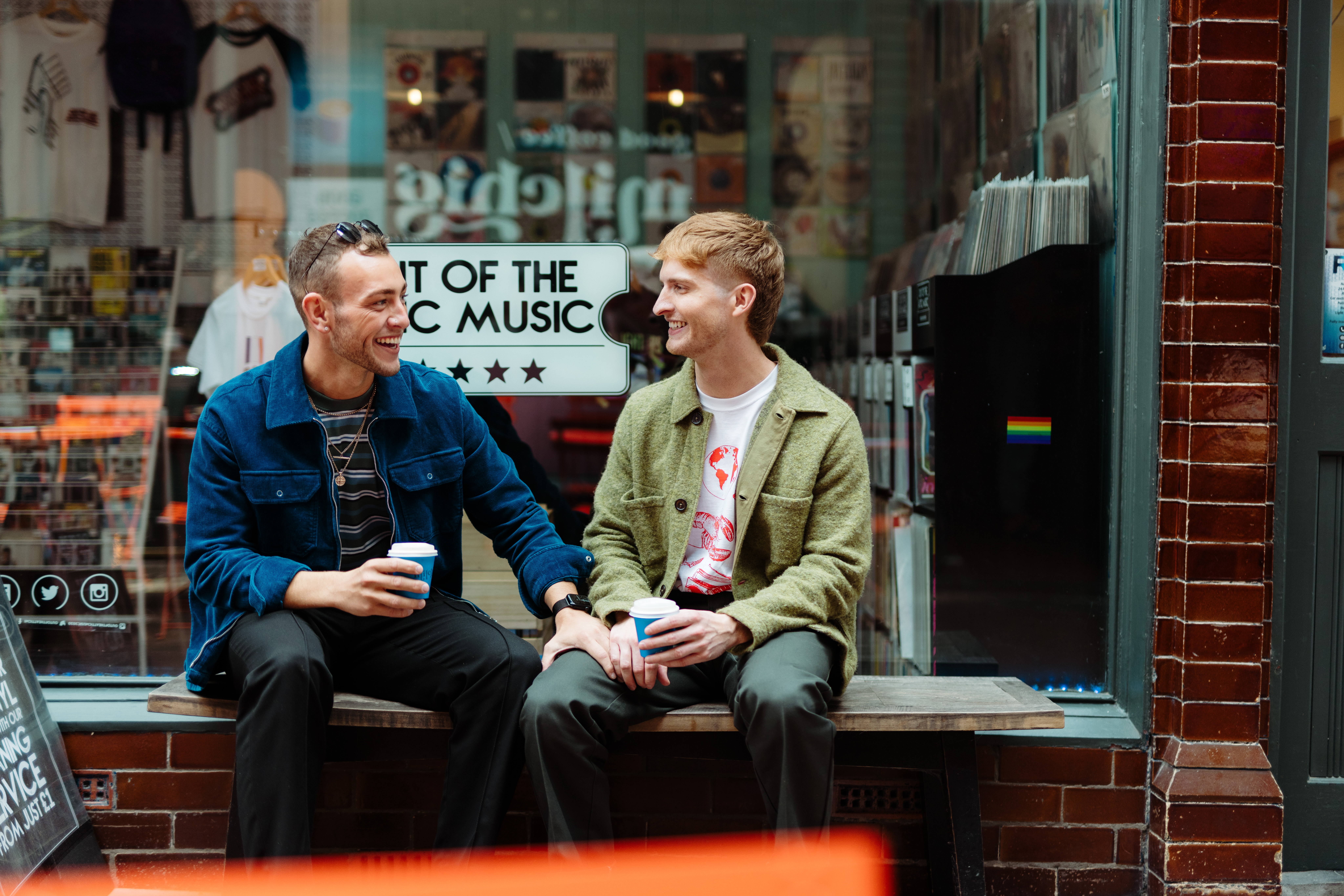 Two men sit with take away drinks in front of a shop