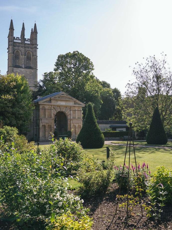 Flower bed in front of a georgian arch and building of Oxford Botanic Garden with church tower behind against blue sky