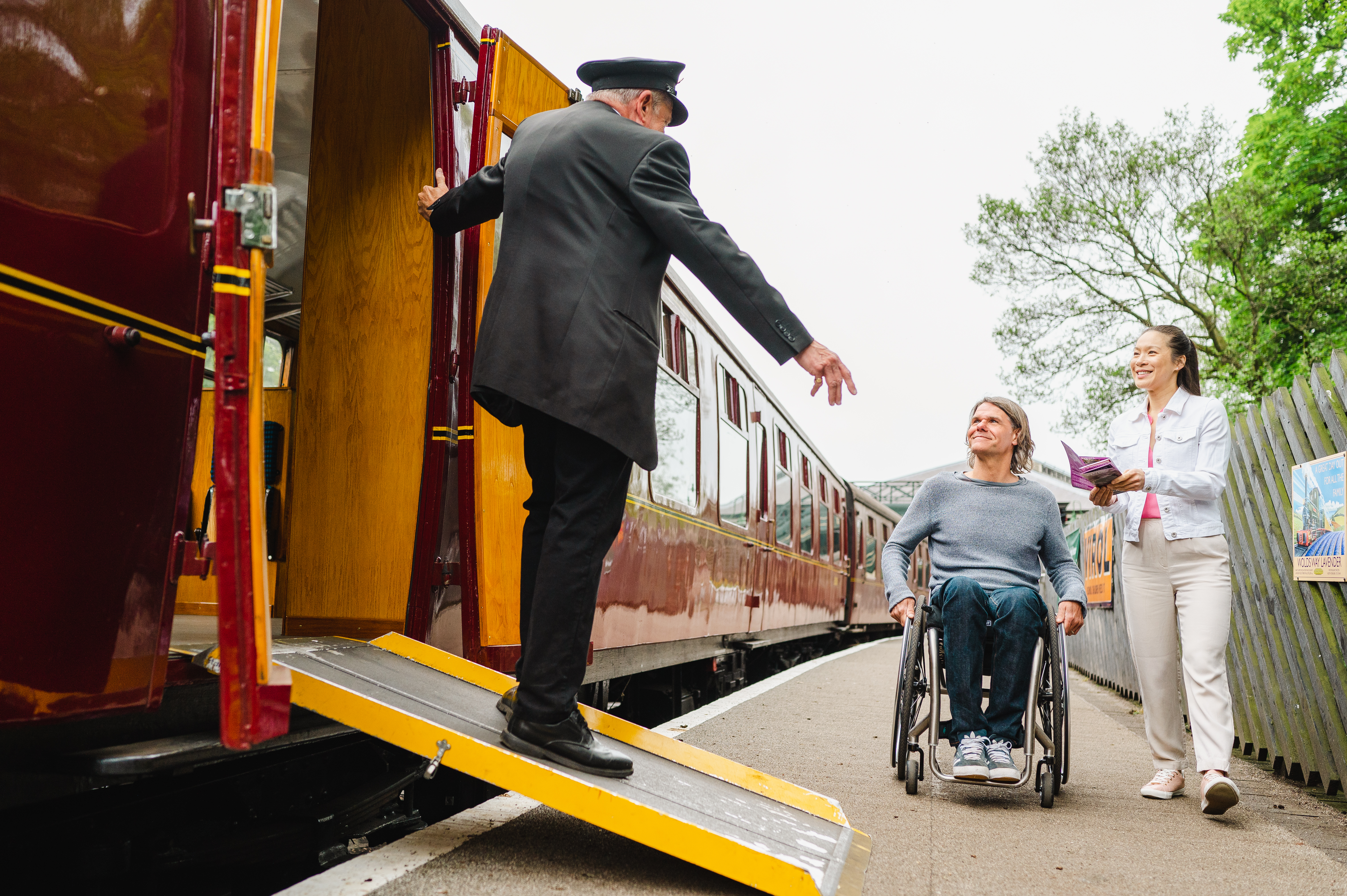Guard stands on a ramps and leans out of heritage train carriage greeting woman and man using a wheelchair