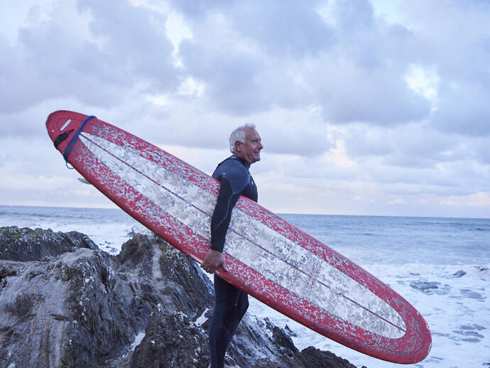 Man carrying a surfboard, standing on the rocks looking out to sea