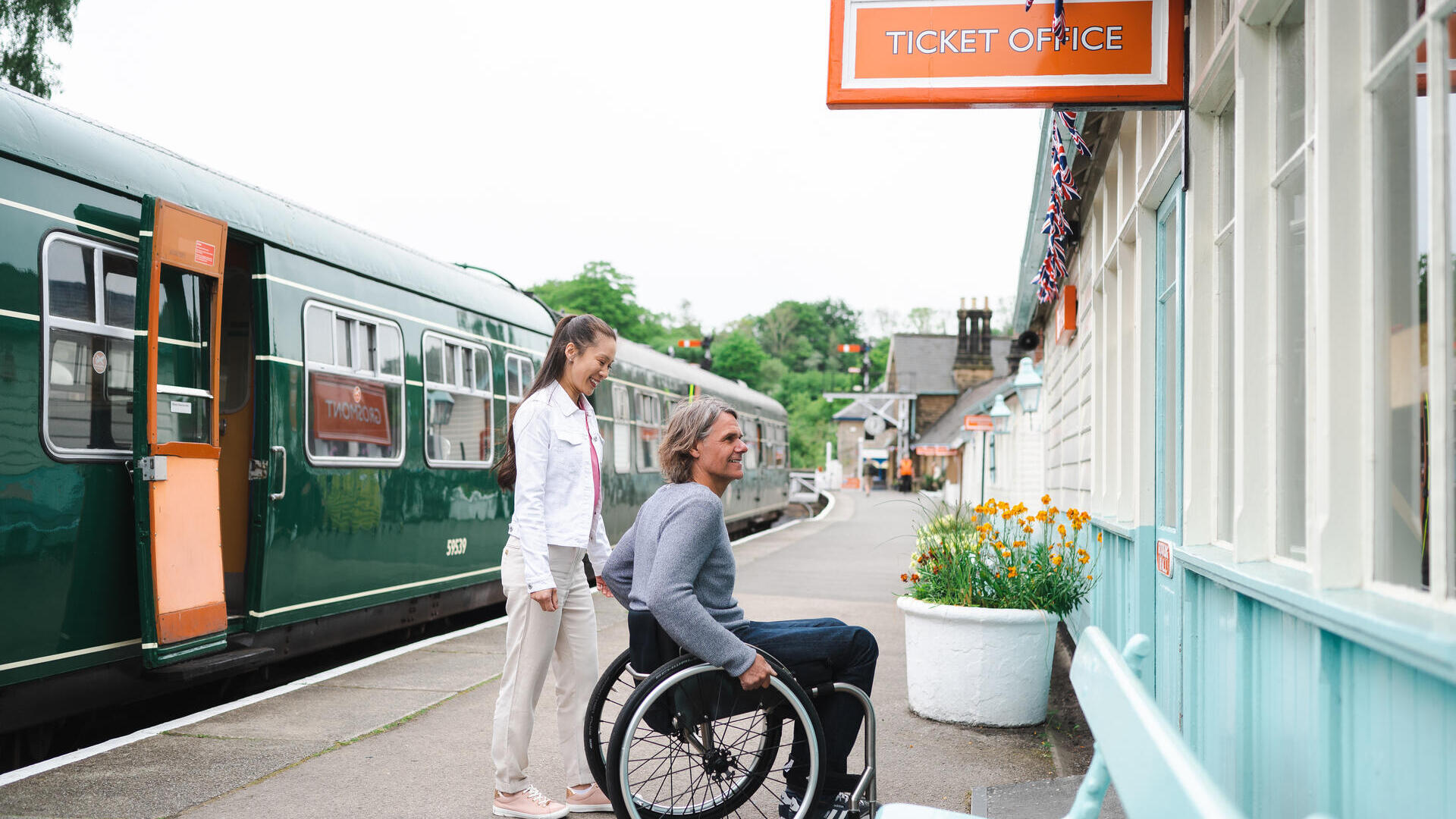 Man using a wheelchair and woman about to go into the ticket office at a station.